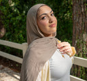 Woman wearing a beige hijab and white top outdoors with greenery in the background
