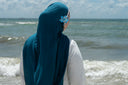Person wearing a blue headscarf and white clothing standing on a beach with waves and sky in the background.
